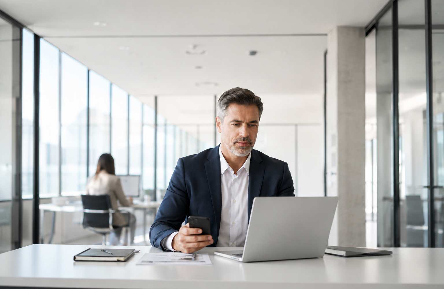 Geschäftsmann prüft eine Nachricht am Smartphone und arbeitet am Laptop in einem hellen modernen Büro als Symbol für CEO-Fraud und Business-E-Mail-Compromise.