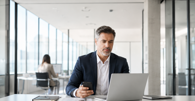 Geschäftsmann prüft eine Nachricht am Smartphone und arbeitet am Laptop in einem hellen modernen Büro als Symbol für CEO-Fraud und Business-E-Mail-Compromise.